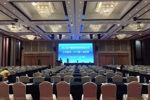 Un salón de conferencias con filas de mesas y sillas, una pantalla grande al frente y candelabros en el techo.