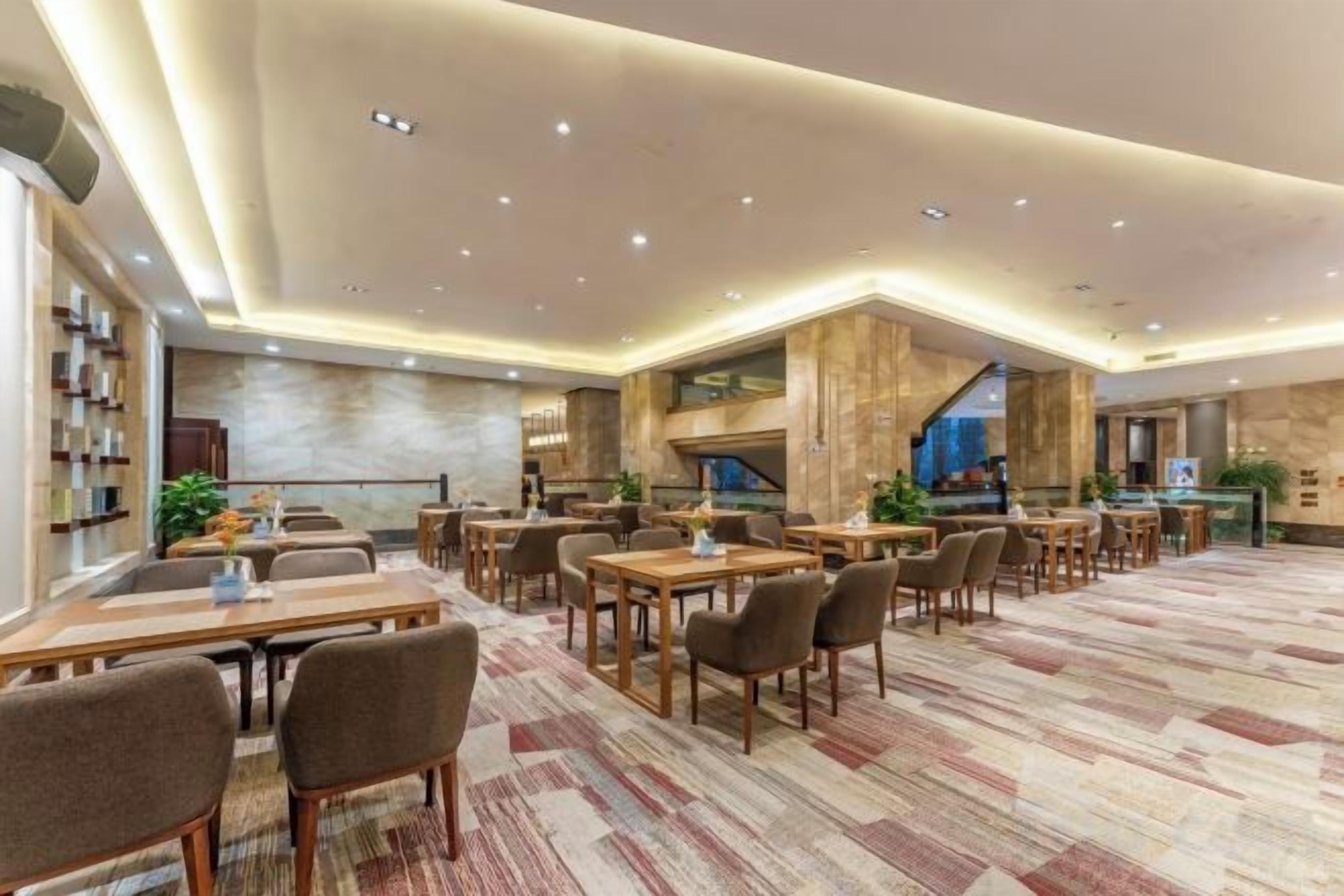 A spacious hotel dining area with multiple tables and chairs, a patterned carpet, and a modern ceiling design.