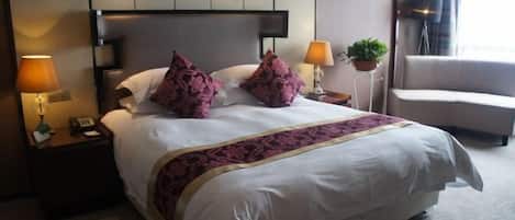 A hotel room with a large bed, two bedside lamps, a nightstand, a potted plant, and a window with curtains.
