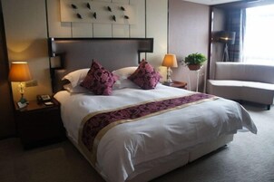 Habitación de hotel con una cama grande, dos lámparas de noche, una mesita de noche, una planta en maceta y una ventana con cortinas.