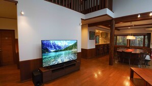 A spacious living area with a large flat-screen TV, wooden flooring, a dining table with chairs, and a kitchen in the background. - Villa Shizen Hakuba (Hakuba)