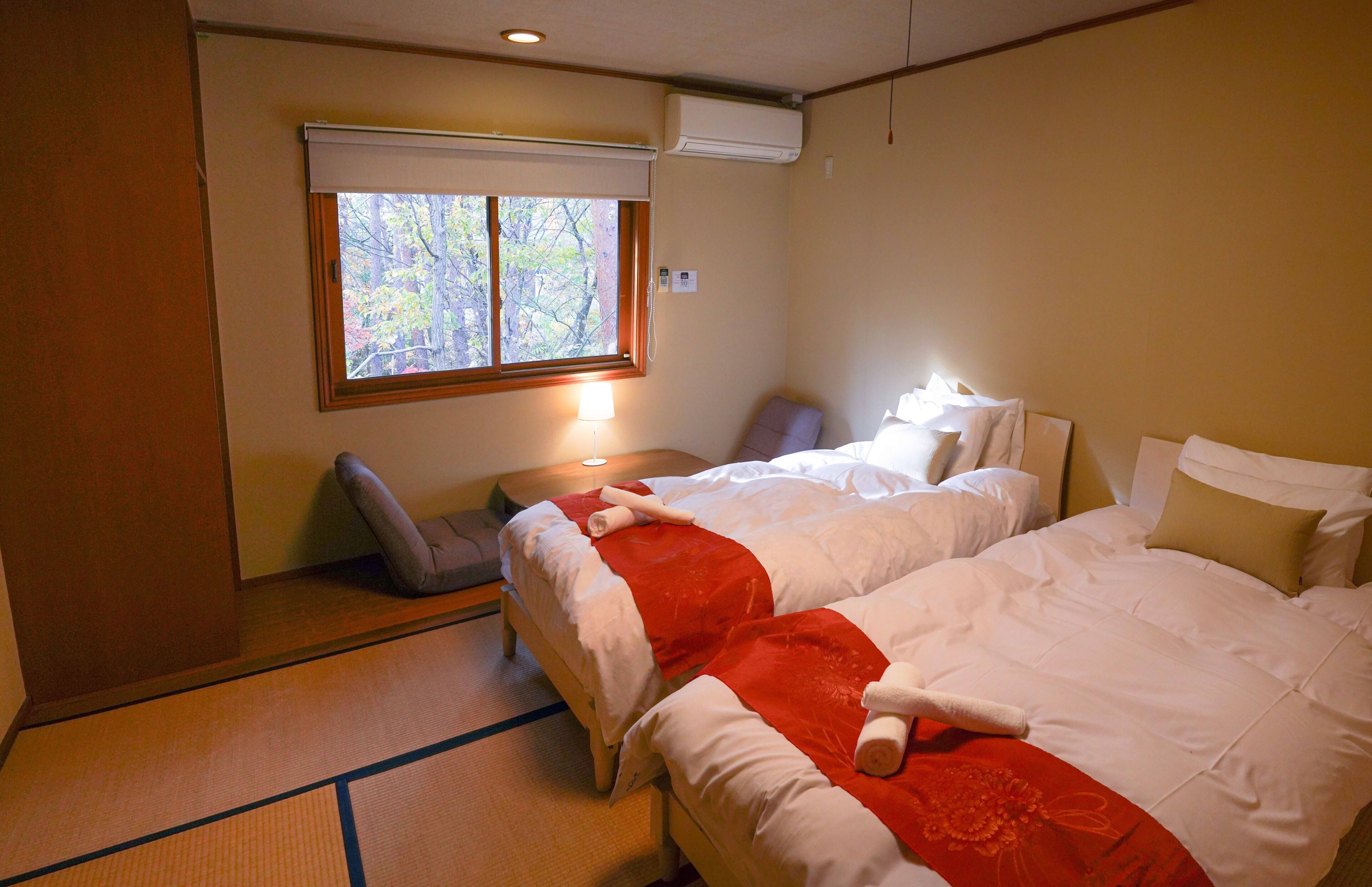 A hotel room with two beds, a window with a view of trees, a bedside table with a lamp, and a chair.