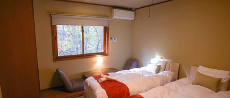 A hotel room with two beds, a window with a view of trees, a bedside table with a lamp, and a chair.