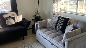 Uma sala de estar com um sofá, uma mesa pequena com abajur e uma janela com vista para o exterior.