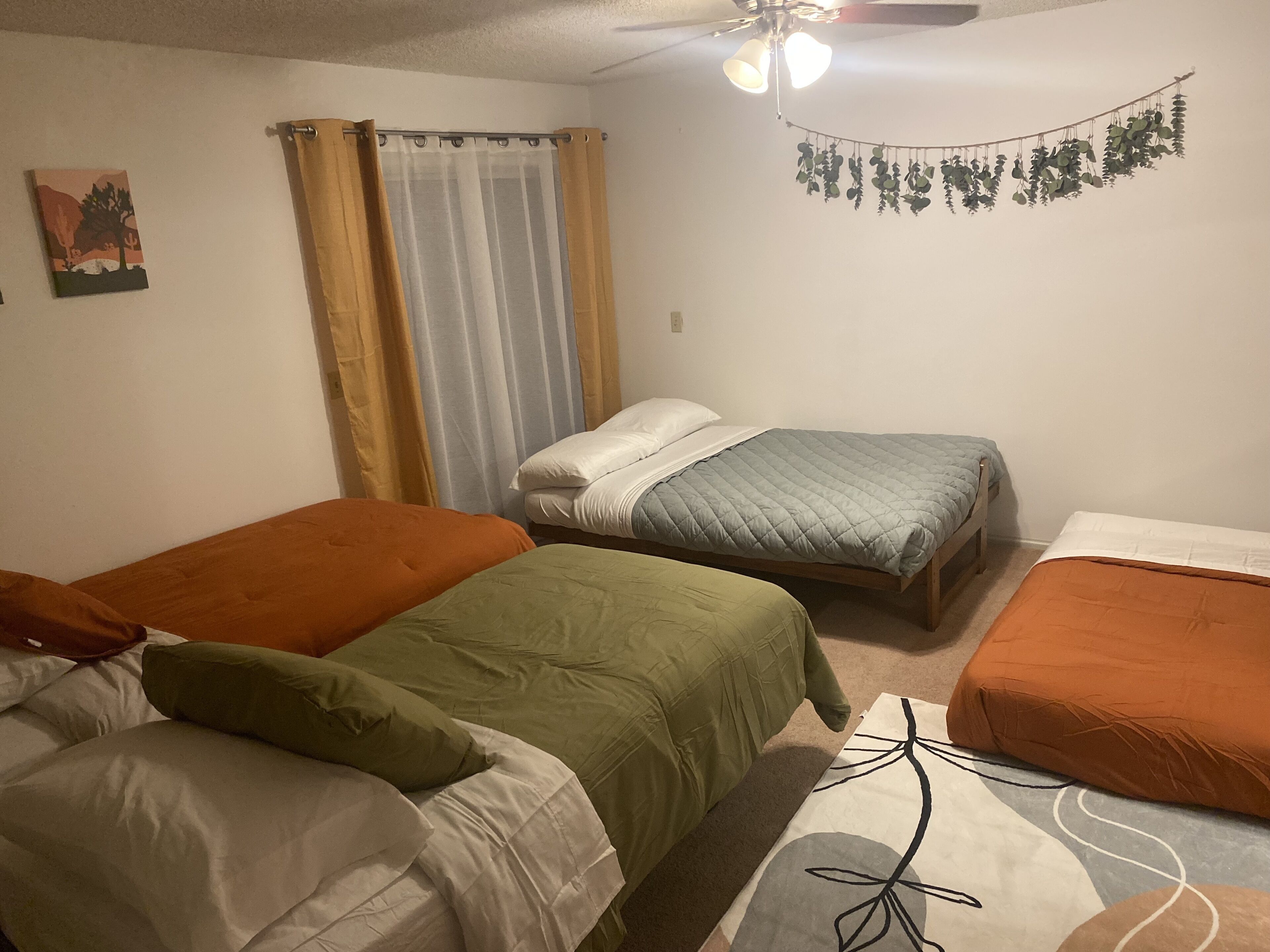 A room with three beds, each with different colored bedding, a ceiling fan, a wall-mounted plant, and a painting on the wall.