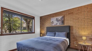 Een slaapkamer met een groot bed, een bakstenen muur, een raam en een tafeltje met een lamp.