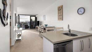 A modern kitchen with a dishwasher, a sink, and a dining area with chairs and a table. - The Lookout - 1-Bedroom Apartment with Stunning Views, Pool & Gym Access, Secure Parking, and Prime Mount Maunganui Location (Mount Maunganui)