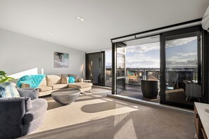 En moderne stue med en stor skydedør, der fører til en balkon, en beige sofa, et sofabord og udsigt over byens skyline.