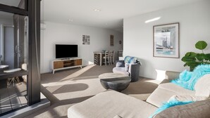 A modern living room with a sofa, a coffee table, a TV, and a dining area with chairs and a table. - The Lookout - 1-Bedroom Apartment with Stunning Views, Pool & Gym Access, Secure Parking, and Prime Mount Maunganui Location (Mount Maunganui)