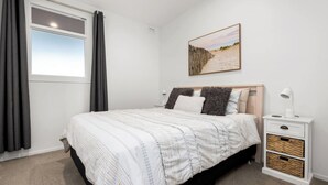 A bedroom with a bed, a nightstand, a lamp, a picture on the wall, and a window with curtains. - The Lookout - 1-Bedroom Apartment with Stunning Views, Pool & Gym Access, Secure Parking, and Prime Mount Maunganui Location (Mount Maunganui)