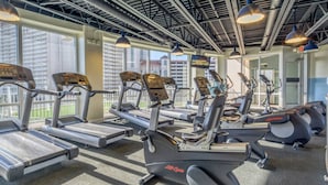 A gym with multiple treadmills and elliptical machines, large windows, and a high ceiling with exposed beams.