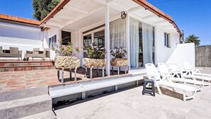A covered outdoor patio with white lounge chairs, a stone planter with plants, and a view of a white building with large windows. - Villa Parralito Ingenio by Interhome (Maspalomas)
