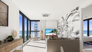 A modern living room with a large window, a sofa, a TV, and a view of the ocean. - B-901 Corner apartment with stunning beach view (Cupecoy)