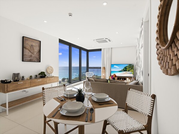 A dining area with a view of the sea, a wooden dining table, white chairs, and a modern kitchen with a wooden cabinet.