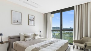 Ein modernes Schlafzimmer mit einem großen Bett, zwei gerahmten Bildern, einem Nachttisch, einem Sessel und Blick auf eine Landschaft durch ein großes Fenster.