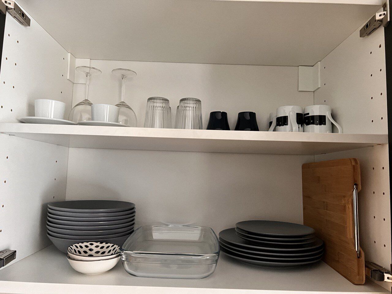 A kitchen shelf with various dishes, glasses, and a cutting board.
