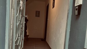 Um corredor com porta de metal, interfone fixado na parede e piso revestido de azulejos.