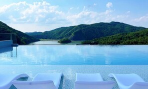 A swimming pool with lounge chairs, surrounded by lush green hills and a clear sky.