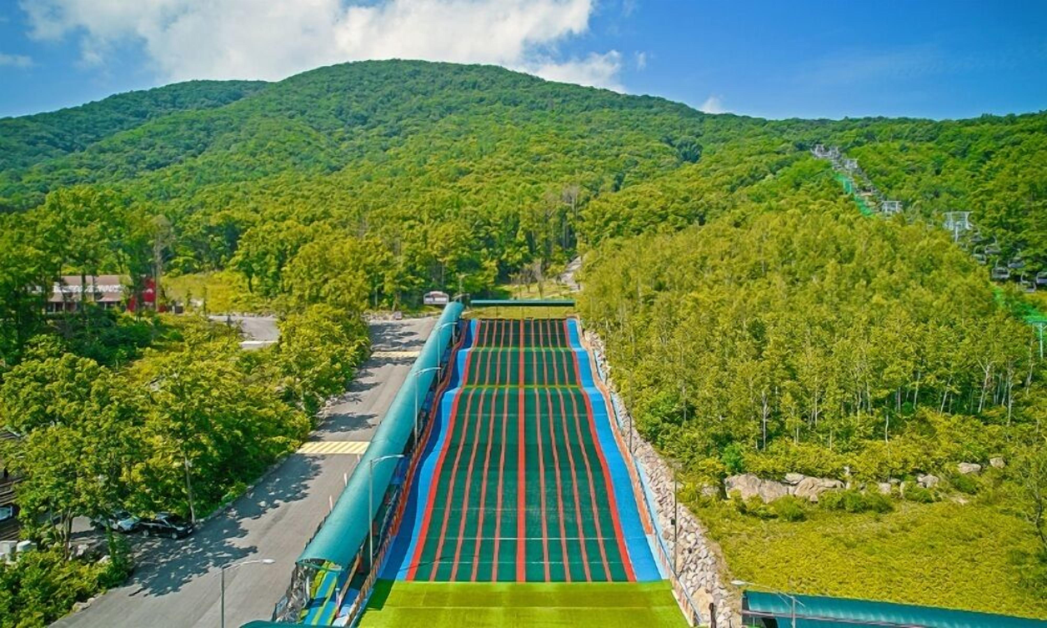 Una estructura de estacionamiento grande, de varios niveles, con techo verde, rodeada de abundante vegetación y una montaña al fondo.