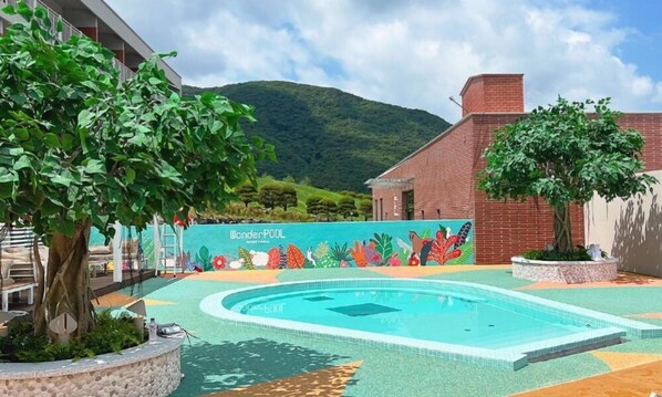 A swimming pool with a mural, surrounded by greenery and a mountain in the background.