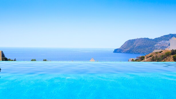 Une magnifique piscine à débordement d’un bleu profond, offrant une vue sur la mer et sur une montagne en arrière-plan.