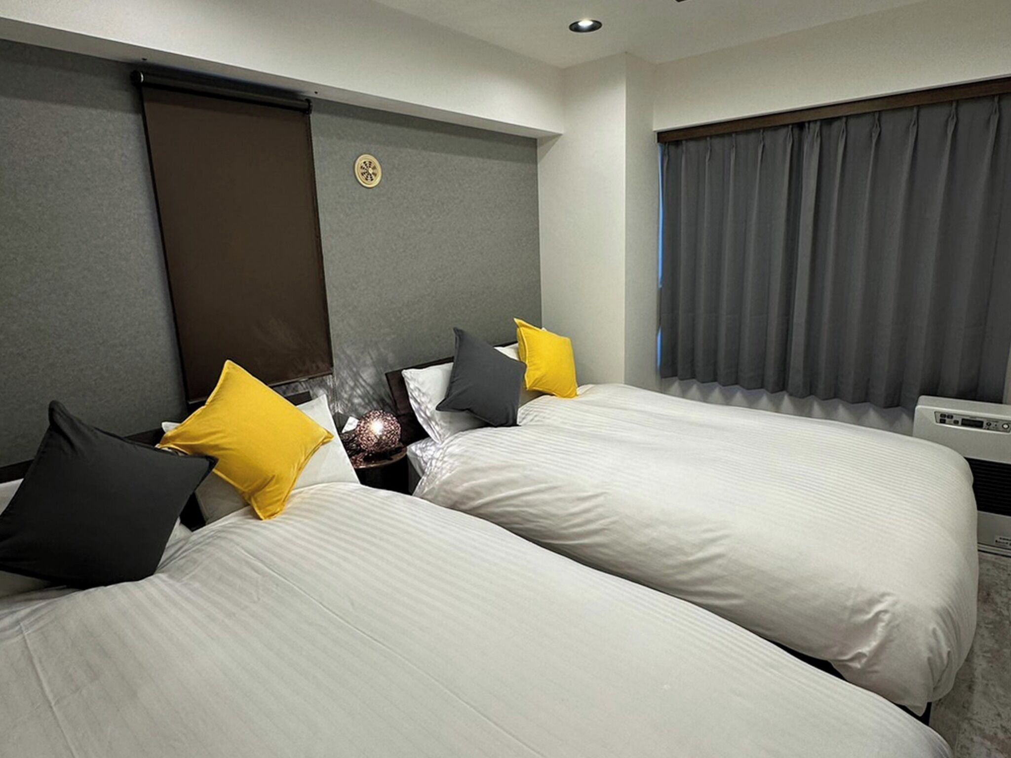 A hotel room with two beds, a bedside table, a lamp, and a window with curtains.