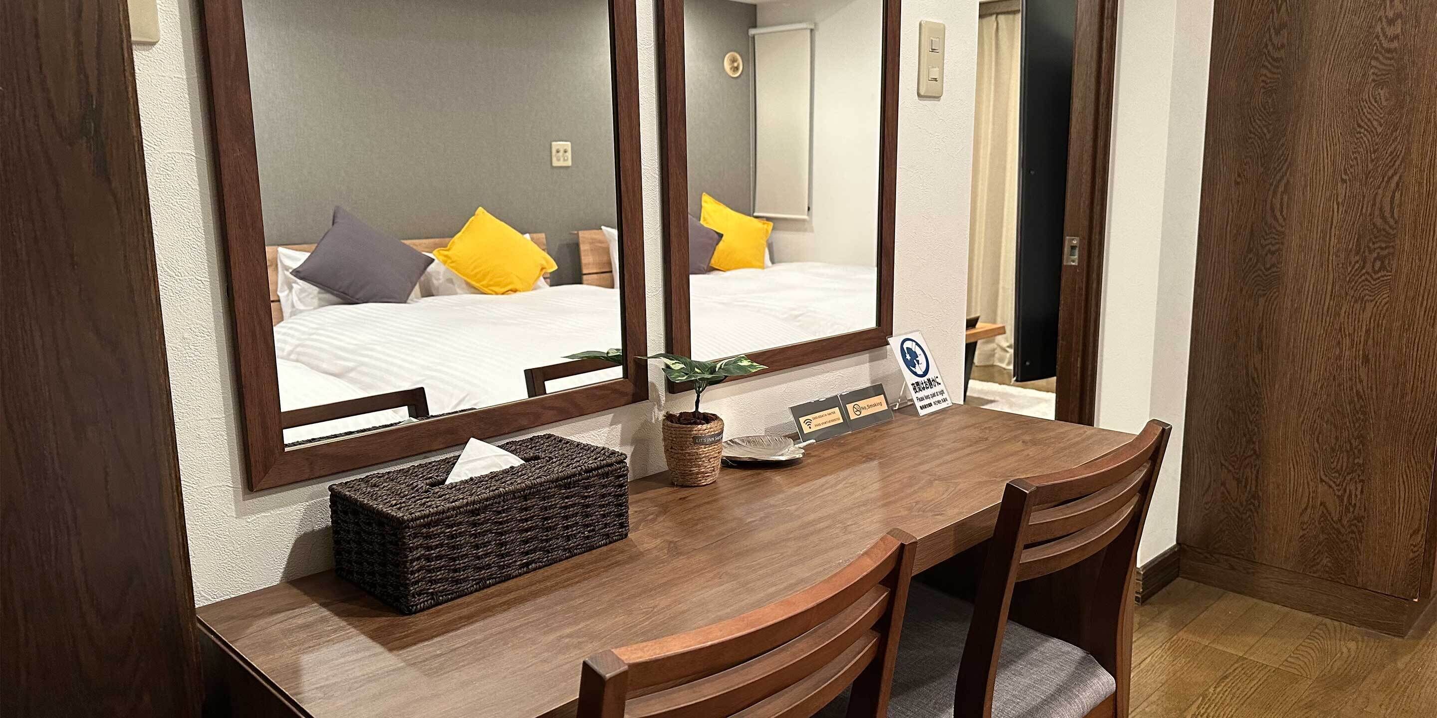 Quarto de hotel com escrivaninha de madeira, duas cadeiras, um espelho grande e uma cama com almofadas amarelas e cinzentas.