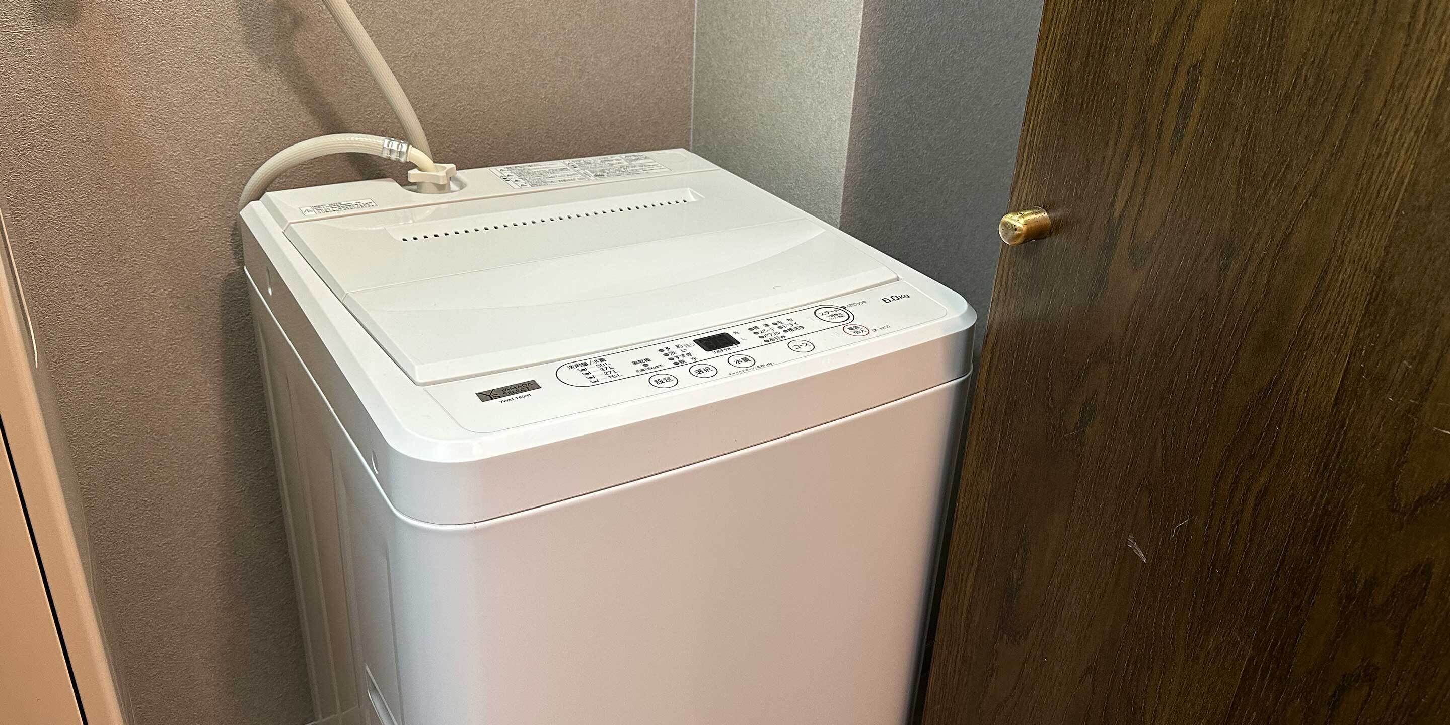 A compact, white, wall-mounted washing machine with a control panel, a power cord, and a door with a brass handle.