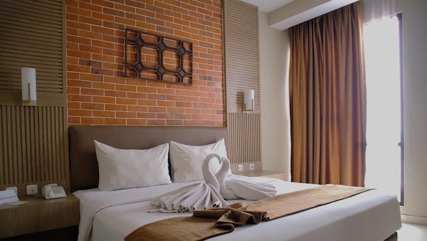 A hotel room with a brick wall, a bed with white linens, a brown throw blanket, and a heart-shaped towel arrangement. - Lynn Hotel Mojokerto (Mojokerto)
