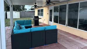 Un patio cubierto con un sofá de mimbre oscuro, una parrilla y un ventilador de techo.