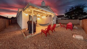 Un espace extérieur accueillant, avec un hamac, des chaises rouges et des guirlandes lumineuses, le tout sous un ciel au coucher du soleil.