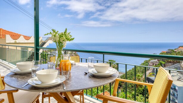 Une table ronde avec de l’assiette, des bols et des verres blancs, un pichet de jus d’orange et un vase rempli de fleurs blanches, le tout placé sur un balcon donnant sur la mer.