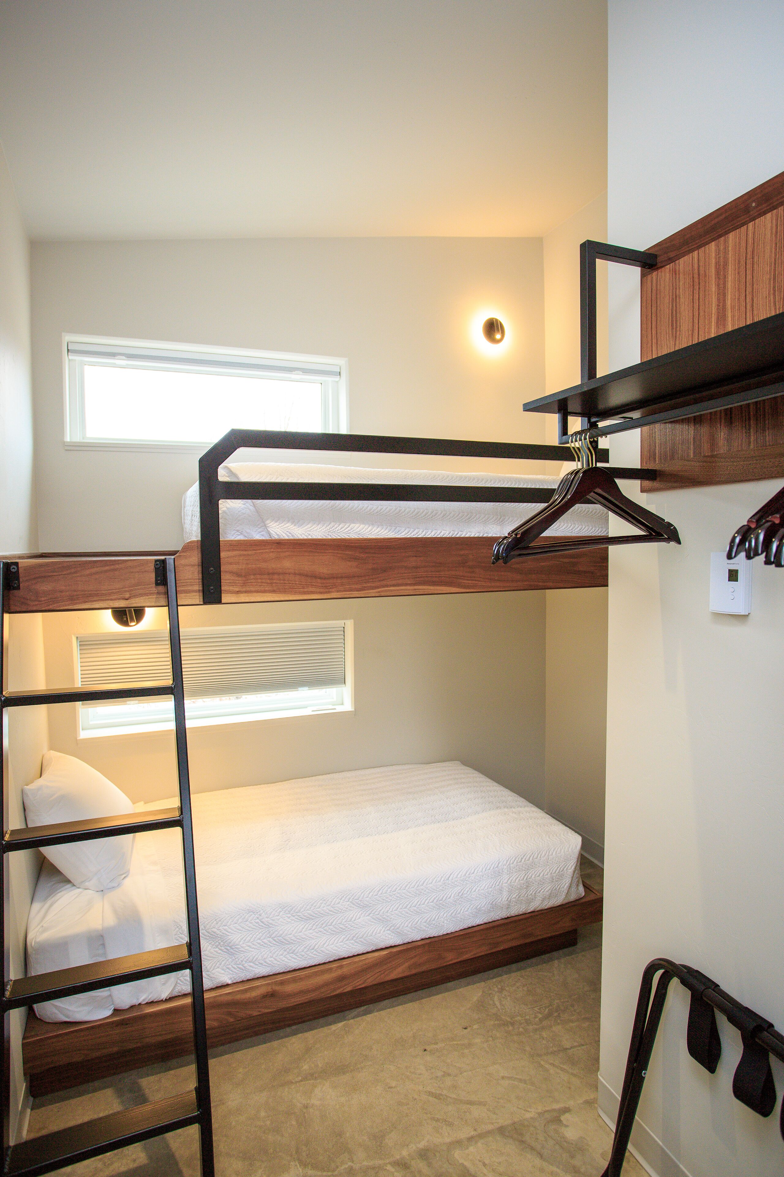 A bunk bed room with a wooden ladder, a hanging coat rack, and a window with blinds.