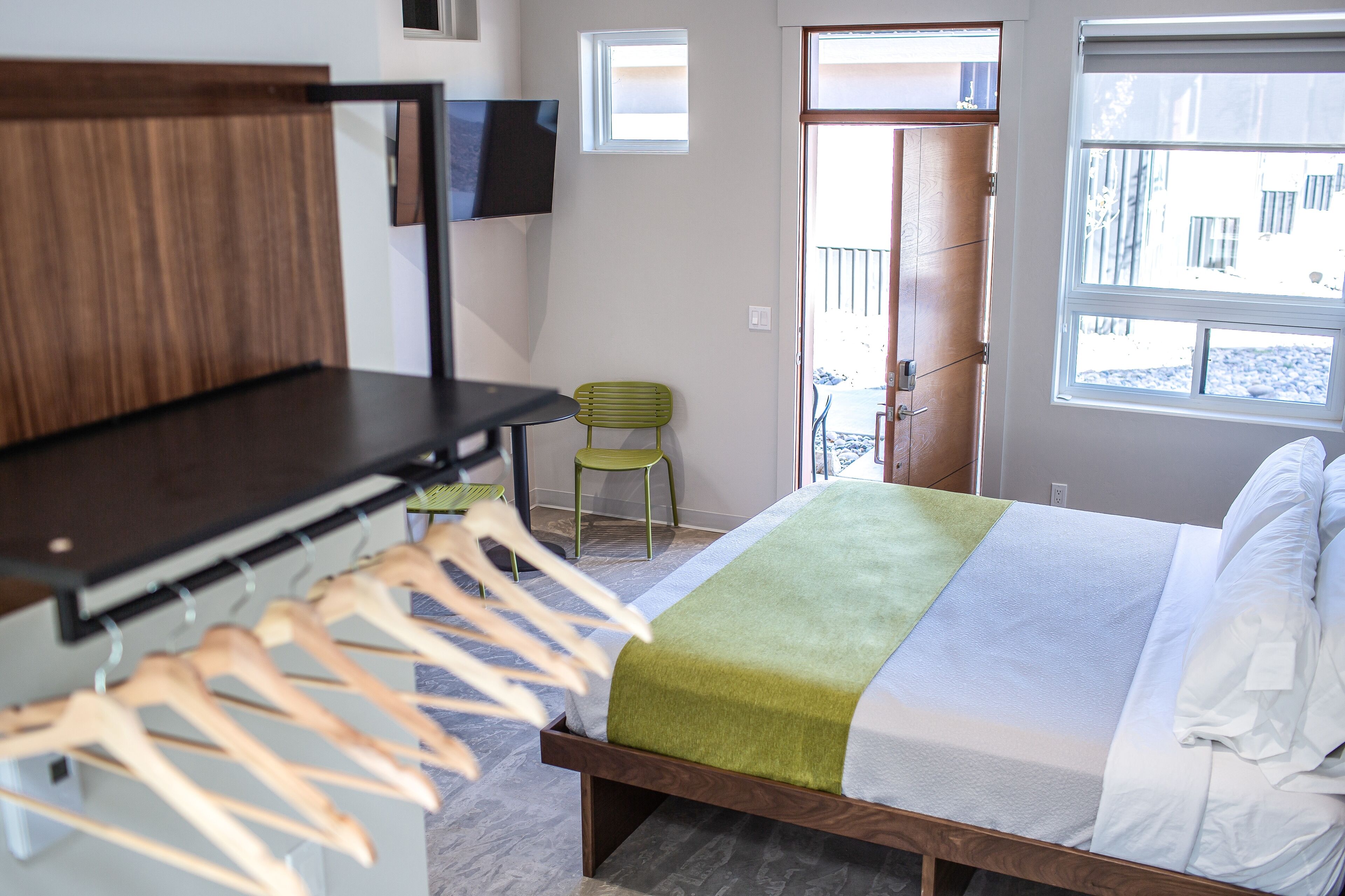 Une chambre avec un lit, une tête de lit en bois, un téléviseur fixé au mur, un fauteuil vert et une fenêtre donnant sur l’extérieur.