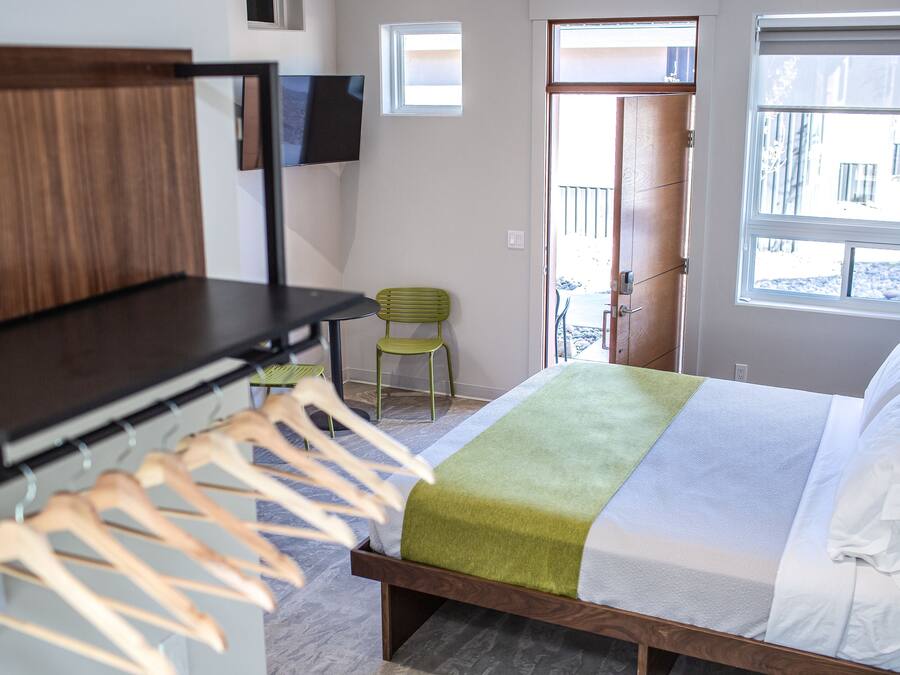 Une chambre avec un lit, une tête de lit en bois, un téléviseur fixé au mur, un fauteuil vert et une fenêtre donnant sur l’extérieur.