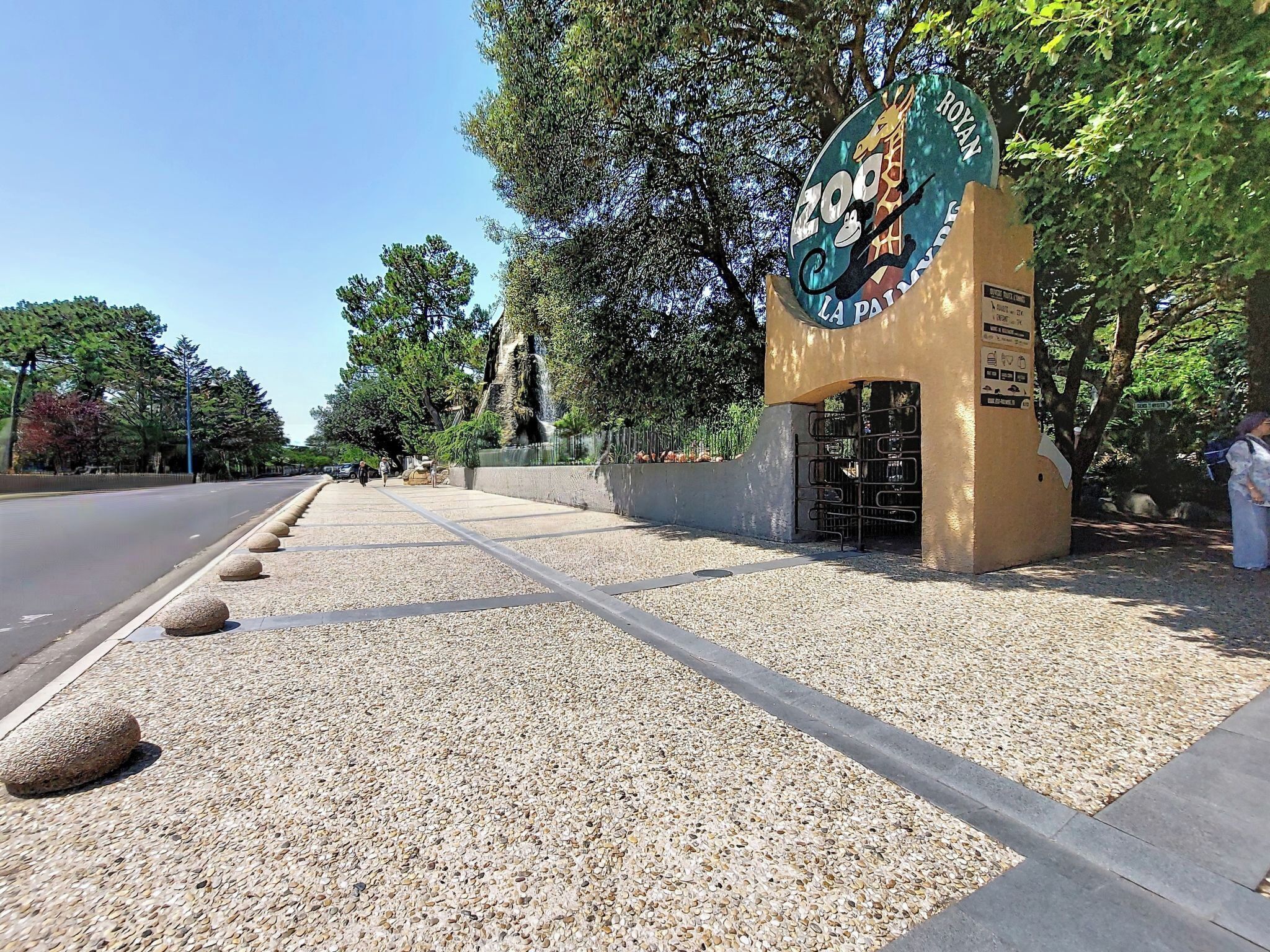 L’entrée d’un zoo, avec un logo représentant une girafe et un singe, un chemin pavé et une zone recouverte de gravier.