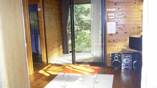 Interior de una cabaña de madera con puerta corrediza de vidrio que da a un balcón, una pequeña mesa y un televisor sobre una estantería.