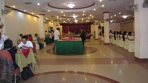 A spacious hall with a central stage, multiple tables with guests, and a buffet setup.