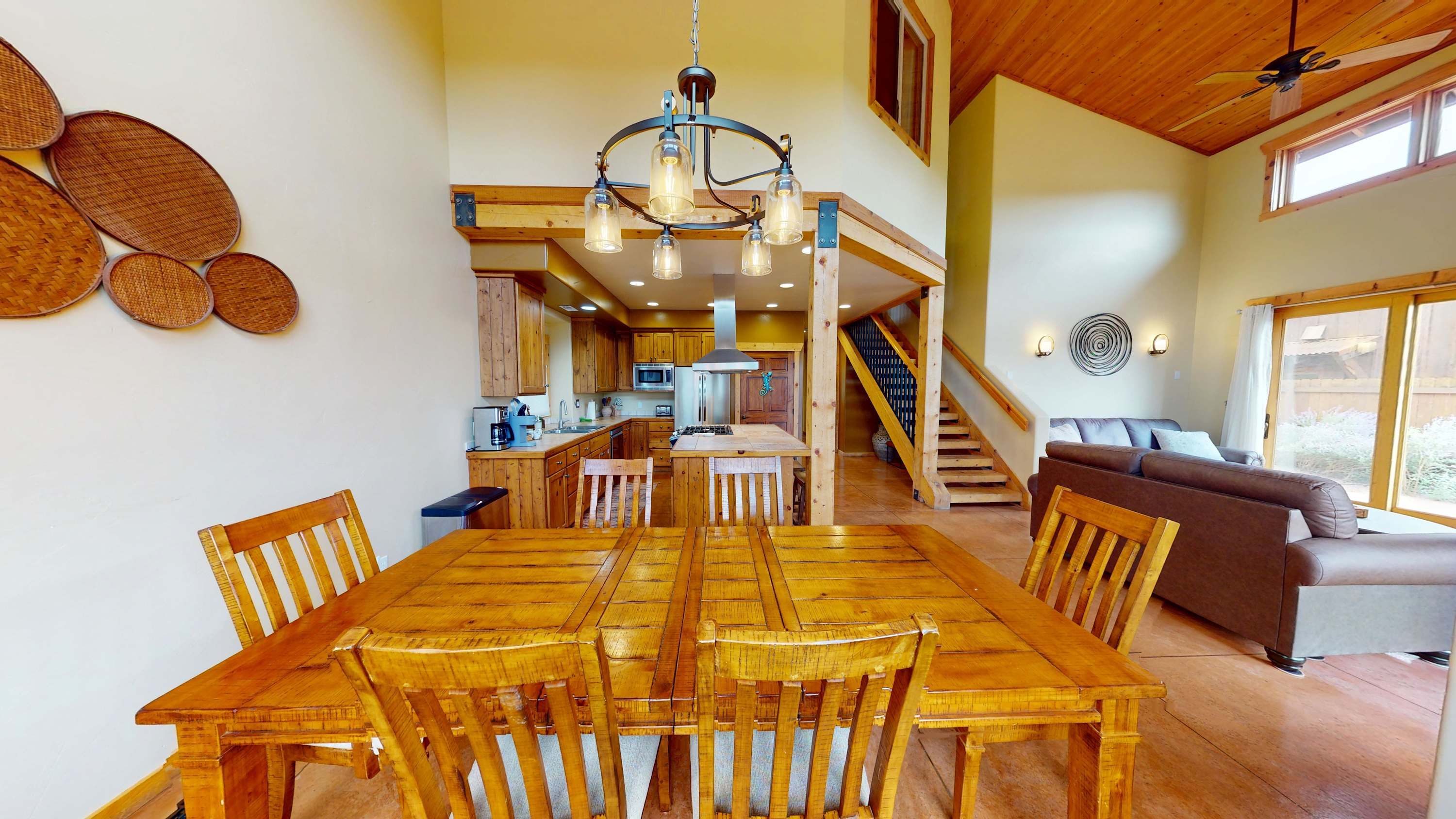 A spacious dining area with a wooden table and chairs, a kitchen in the background, and a staircase leading to an upper level.