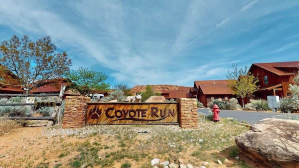 A sign for 'Coyote Run' with a stone wall and wooden gate, surrounded by desert vegetation and buildings with red roofs.