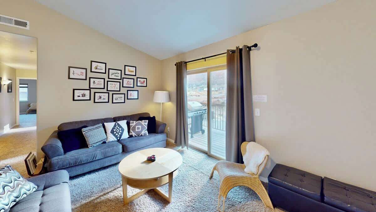 A living room with a sofa, a round coffee table, and a chair. There are framed pictures on the wall and a window with curtains.