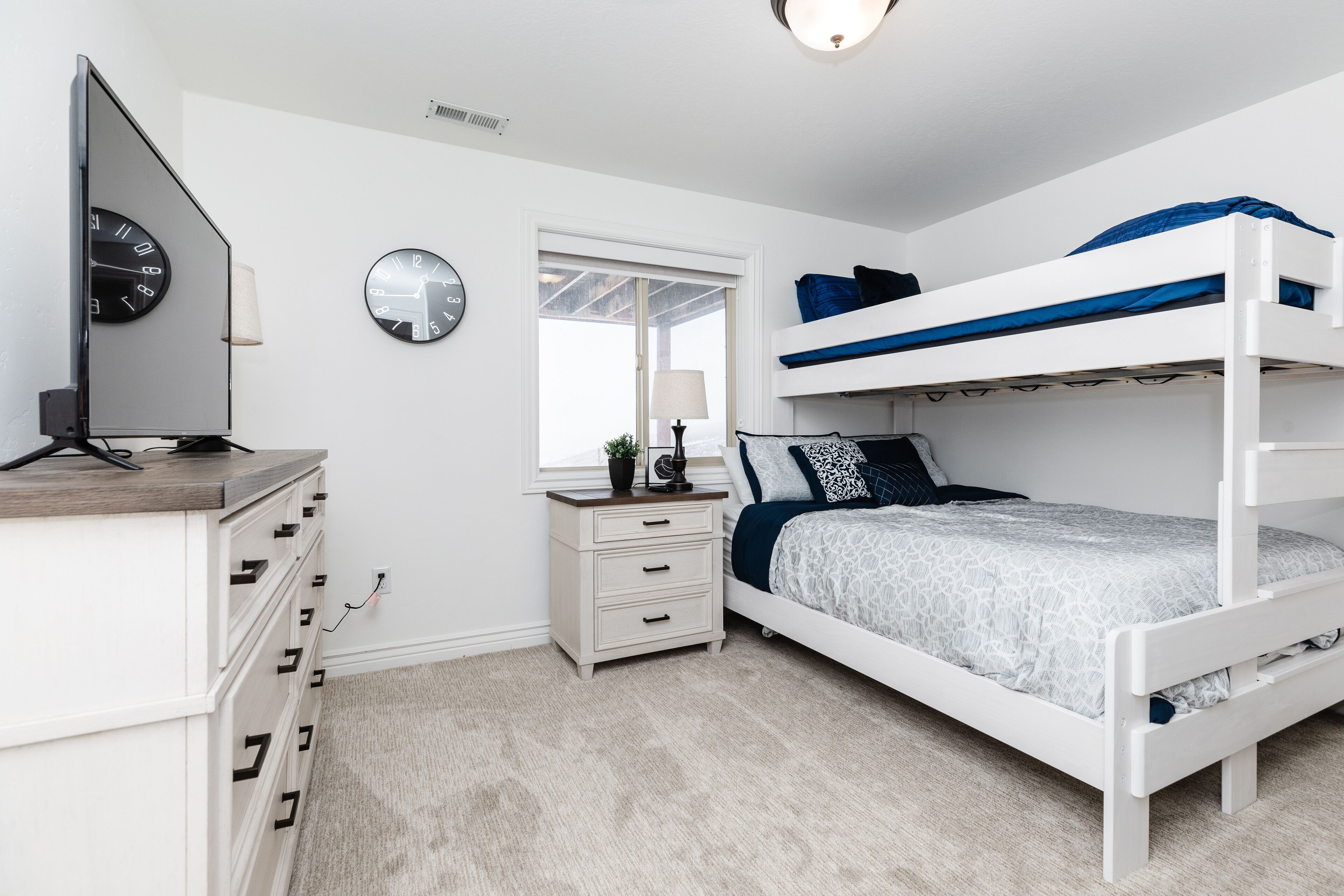 A bedroom with a bunk bed, a desk with a computer, a dresser, two wall clocks, and a window.
