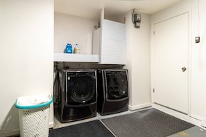 A laundry area with two front-loading washers, a trash can, and a white door. - The Lakehouse at Pinellas Park (Pinellas Park)