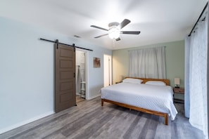 A bedroom with a bed, a ceiling fan, a door, and a nightstand. - The Lakehouse at Pinellas Park (Pinellas Park)