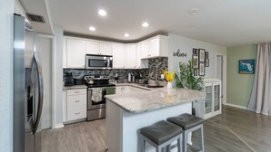 Una cocina moderna con armarios blancos, electrodomésticos de acero inoxidable, una isla grande con taburetes y un cartel con la palabra 'welcome'.