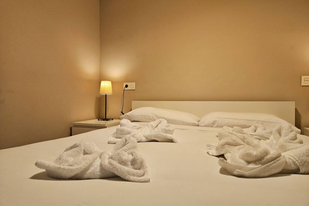 Habitación de hotel con cama, mesita de noche, lámpara y dos juegos de toallas blancas.