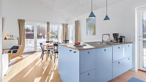 A modern kitchen with a central island, blue pendant lights, and a view of the outdoors through large windows.