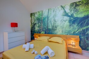 A bedroom with a bed, pillows, towels, a nightstand, a lamp, and a wall mural.