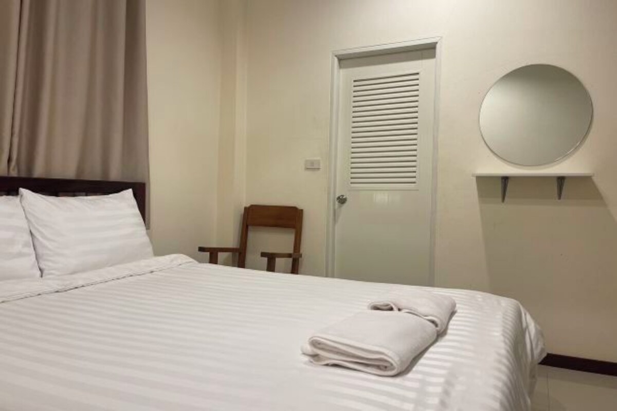 A neatly made bed with white linens, a wooden chair, a door with a window, and a round mirror on the wall.