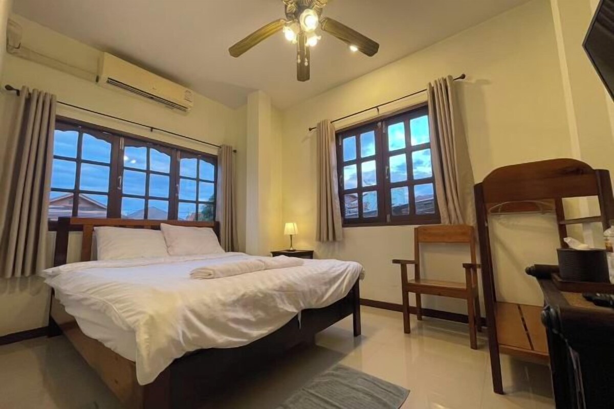 A hotel room with a bed, two windows, a ceiling fan, and a chair.